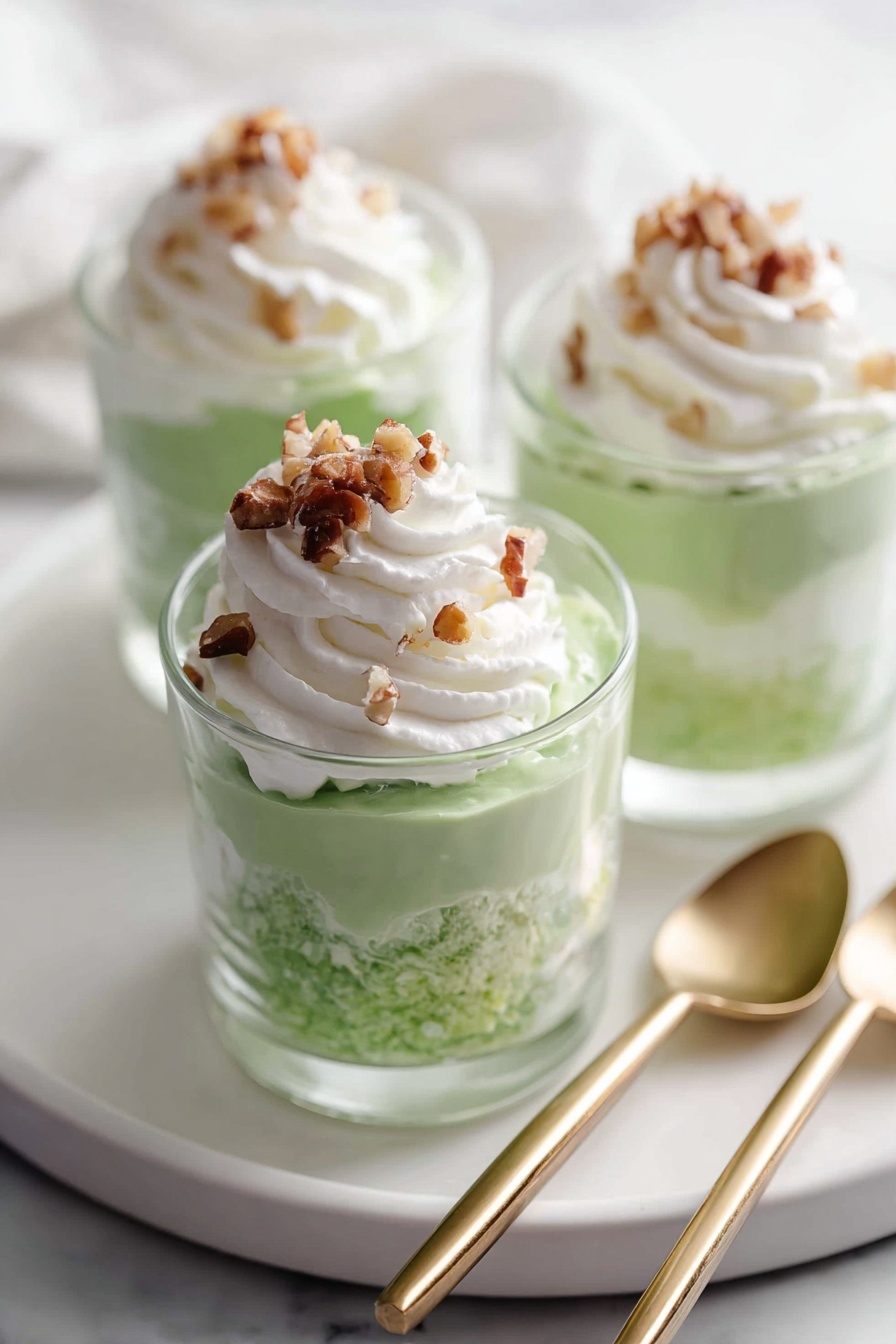 Three clear glass cups each hold a layered green creamy dessert with a fluffy texture. The green layer fills most of the cup, topped with a swirl of white whipped cream. On top of the whipped cream, there are small pieces of chopped brown nuts. The cups are placed on a round white plate with a smooth surface, set on a white marbled background. Two gold-handled spoons rest on the surface next to the plate. Photo taken with an iphone --ar 2:3 --v 7 - Easy Pistachio Watergate Salad, Pistachio Watergate Salad, Watergate Salad with Pistachios, No-Bake Watergate Salad, Retro Pistachio Fruit Salad
