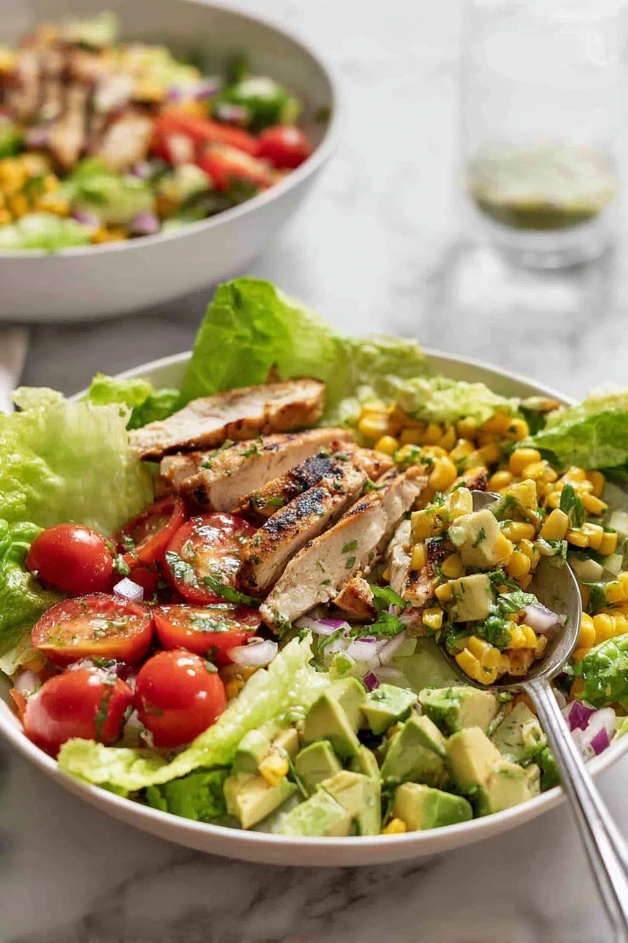A white bowl filled with a fresh salad showing three main layers: the bottom layer is large green lettuce leaves with a crisp texture, the middle layer has ripe cherry tomatoes, yellow corn kernels, chopped avocado, and small pieces of red onion, all mixed together, and the top layer is grilled chicken slices with light brown grill marks and green herbs sprinkled on top. On the right side of the bowl, a spoon holds a mixture of diced avocado and corn. In the background, a second white bowl with a similar salad is slightly out of focus, and the surface is a white marbled texture. Photo taken with an iphone --ar 2:3 --v 7 - Mexican Chicken and Avocado Salad, healthy Mexican chicken salad, easy Mexican chicken recipes, vibrant lunch salads, fresh avocado chicken dish