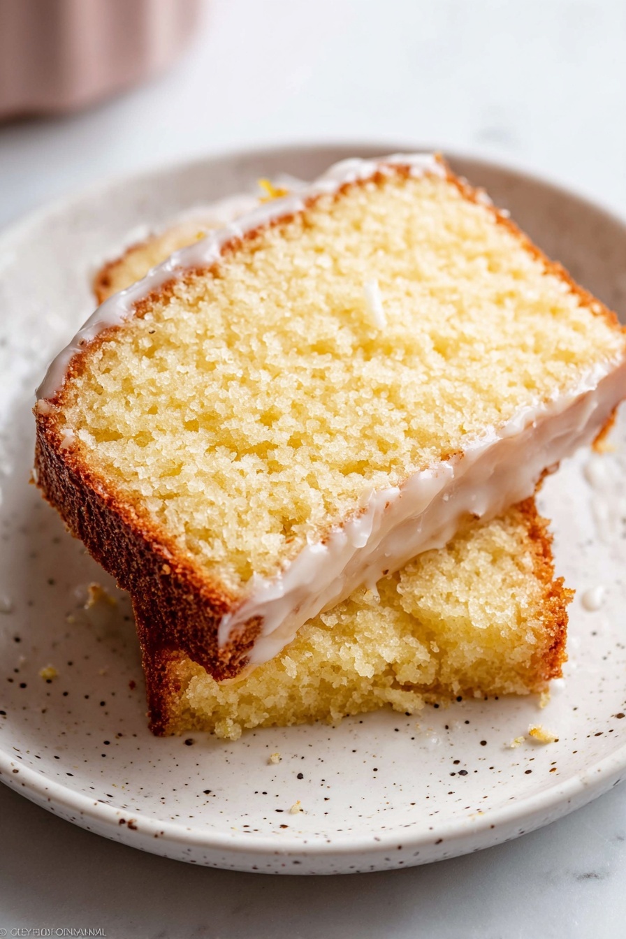 Two slices of yellow cake with a light texture and crumbly edges are stacked on a white plate with small black specks. The top slice has a thin layer of white glaze that partly covers the surface, showing a moist interior. The cake edges are golden brown, with a soft, spongy middle layer that is pale yellow. The plate sits on a white marbled surface, and the image has soft natural light highlighting the texture of the cake. photo taken with an iphone --ar 2:3 --v 7 - Iced Lemon Pound Cake, Lemon Pound Cake, citrus dessert, easy lemon cake, moist pound cake